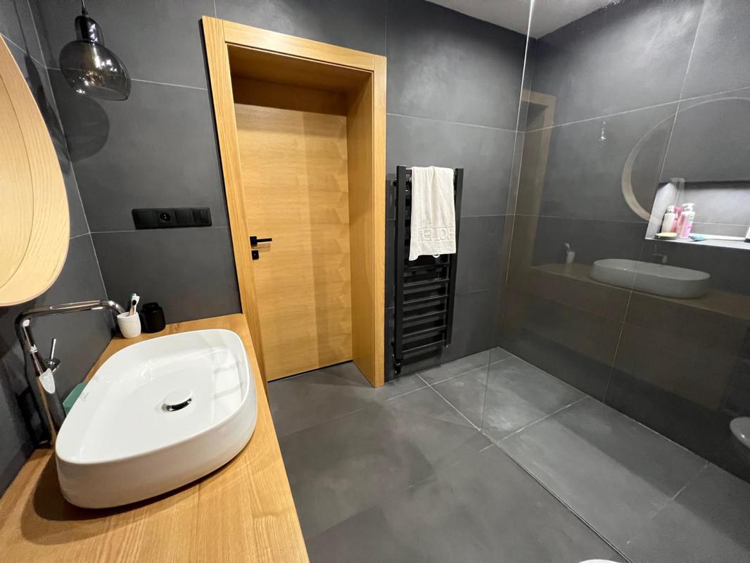 Kombinace dvou odstínů obkladů a dlažby v imitaci betonu vytváří harmonický kontrast mezi světlejšími a tmavšími tóny. Eleganci a jedinečný styl koupelně podtrhují moderní prvky, jako je umyvadlo umístěné na desce a stojánková vodovodní baterie.